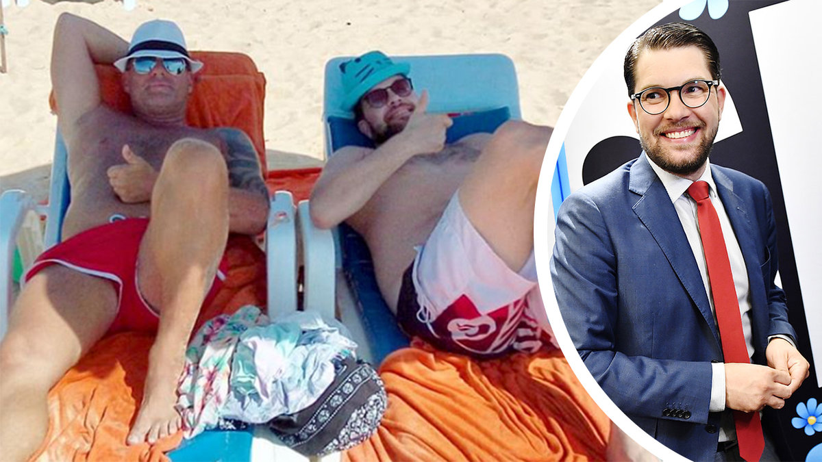 två man i badkläder ligger på varsin solstol på stranden, en man i mörkt hår och blå kostym, med röd slips och svarta runda glasögon