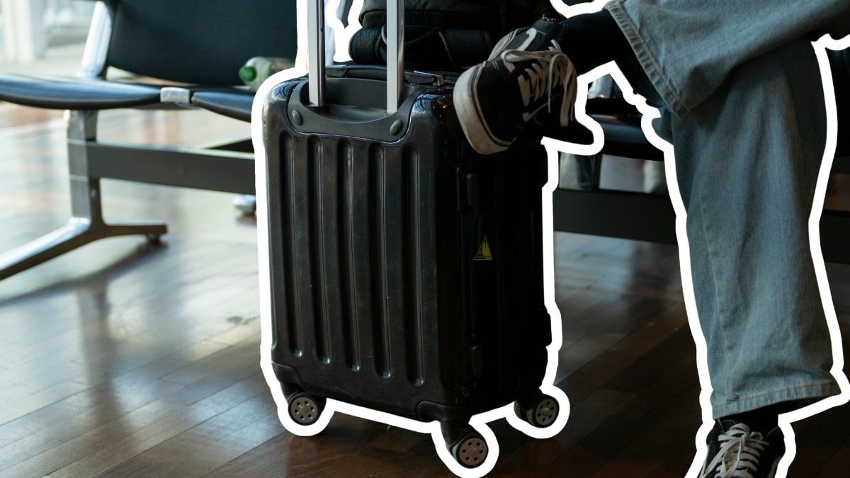 En person sitter på en bänk med en handbagage-resväska bredvid sig.