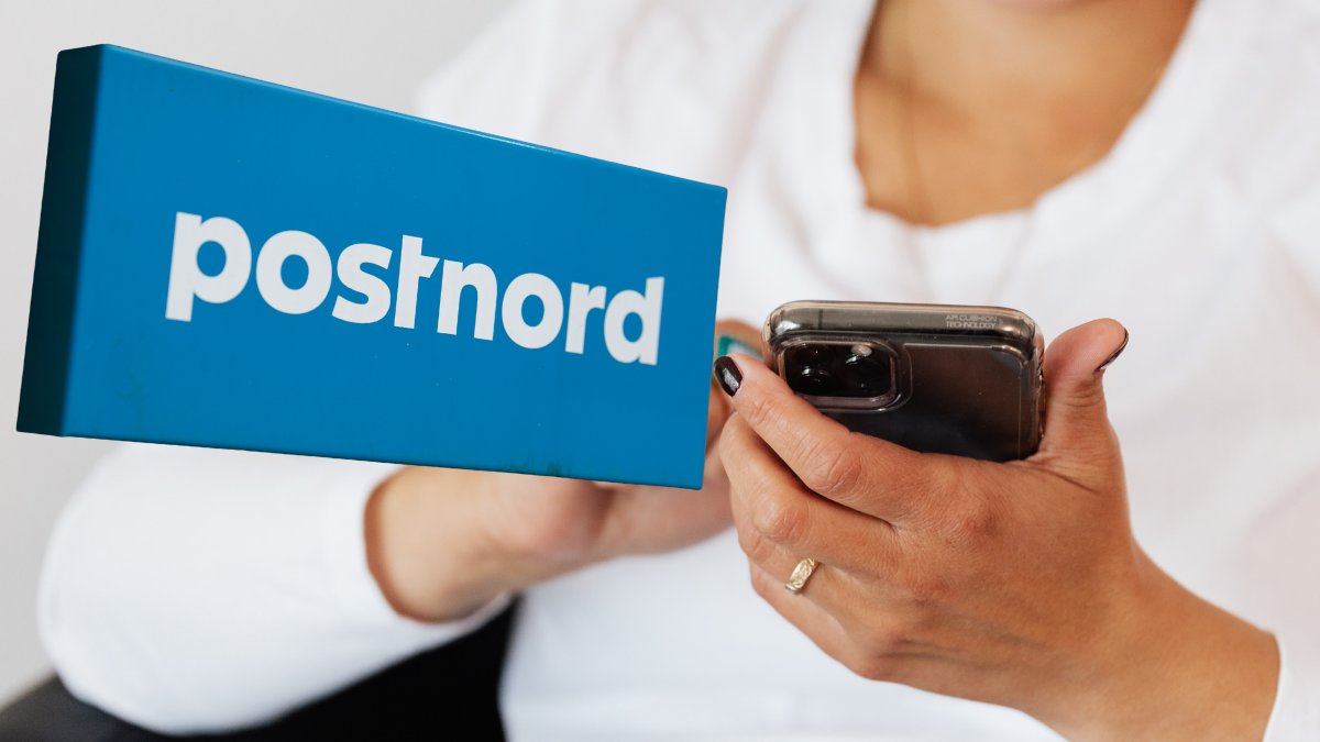En person som håller i en telefon i bakgrunden. En utklippt bild på en Postnord-skylt vänster.