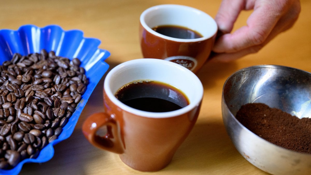 Två kaffekoppar på ett bord. Höger om dem ligger malet kaffe. Vänster om dem ligger kaffebönor.