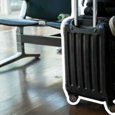 En person sitter på en bänk med en handbagage-resväska bredvid sig.