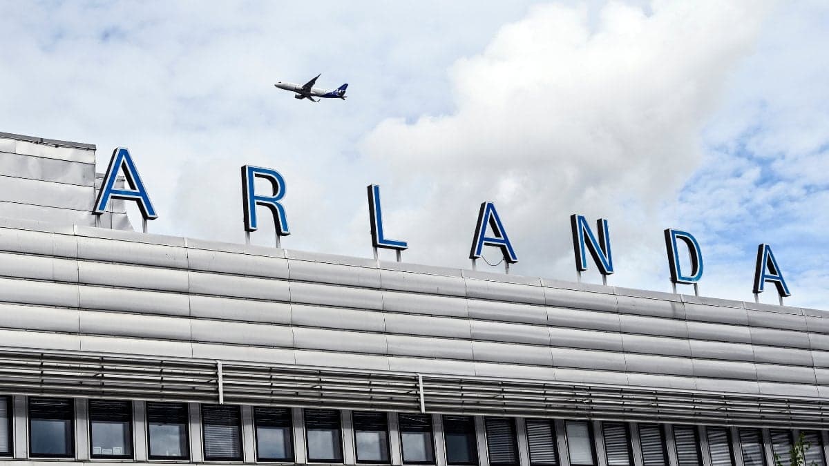Arlanda-skylt vid flygplatsens ingång. I bakgrunden ser man ett flygplan flyga i himlen.