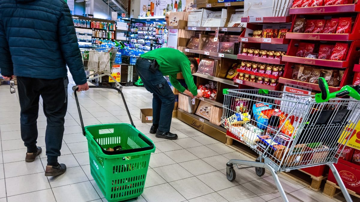 En kund som går i en matbutik med en korg medan en anställd fyller på hyllorna i bakgrunden.